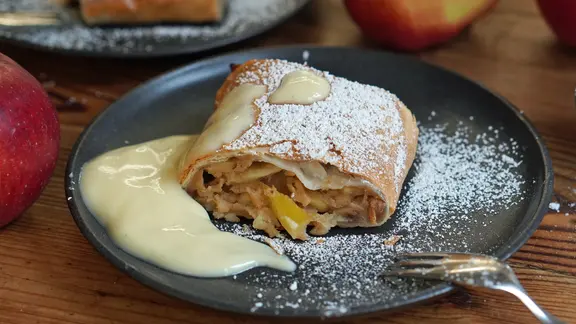 Ein Stück Apfelstrudel mit Puderzucker und Vanillesoße auf einem dunklen Teller